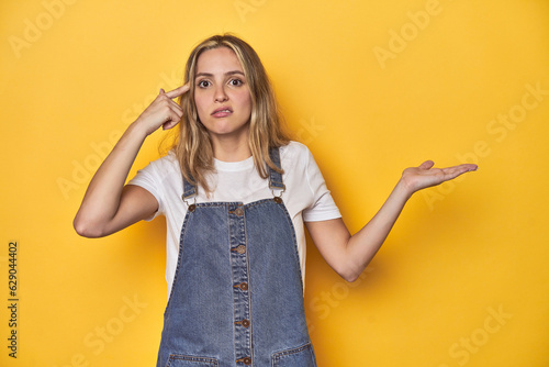 Young blonde Caucasian woman in denim overalls posing on a yellow background, holding and showing a product on hand.