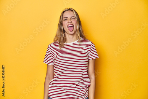 Young blonde Caucasian woman in a red striped t-shirt on a yellow background, funny and friendly sticking out tongue.
