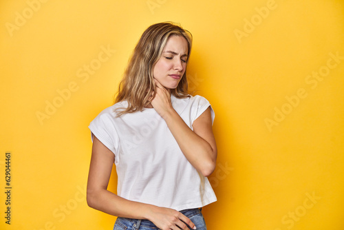 Young blonde Caucasian woman in a white t-shirt on a yellow studio background, suffers pain in throat due a virus or infection. photo
