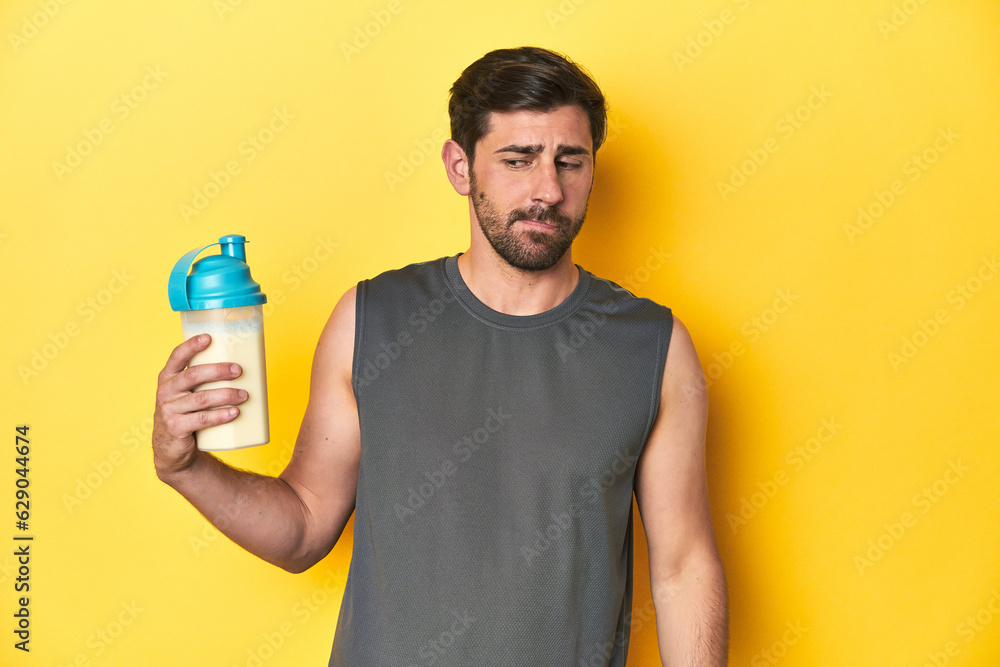 Fit man with protein shake, yellow studio background confused, feels doubtful and unsure. Fit man with protein shake, yellow studio background confused, feels doubtful and unsure.