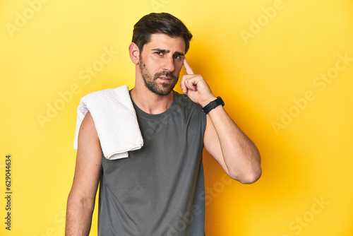 Sporty Caucasian man with towel, studio shot on yellow Sporty Caucasian man with towel, studio shot on yellowpointing temple with finger, thinking, focused on a task.