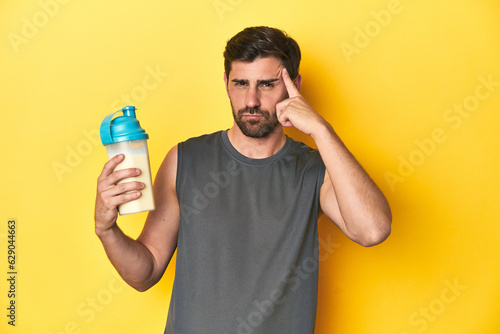 Fit man with protein shake, yellow studio background pointing temple with finger, thinking, focused on a task.