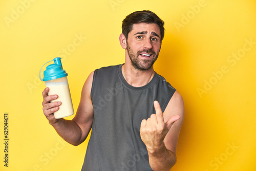 Fit man with protein shake, yellow studio background pointing with finger at you as if inviting come closer.