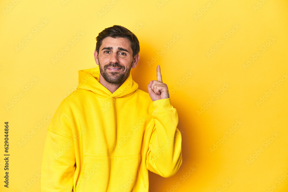 Man in yellow hoodie, monochrome studio backdrop showing number one with finger. Man in yellow hoodie, monochrome studio backdrop showing number one with finger.
