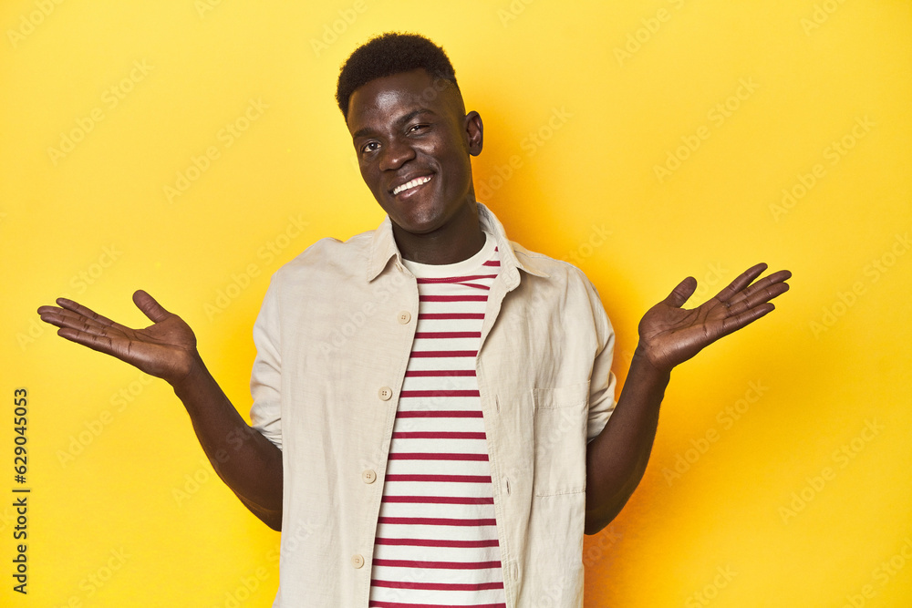 Stylish young African man on vibrant yellow studio background, confused and doubtful shrugging shoulders to hold a copy space. Stylish young African man on vibrant yellow studio background, confused and doubtful shrugging shoulders to hold a copy space.