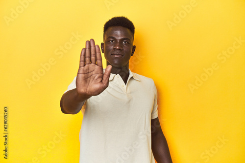 Stylish young African man on vibrant yellow studio background, standing with outstretched hand showing stop sign, preventing you.