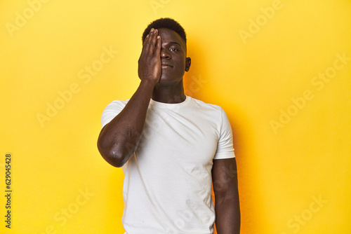 Stylish young African man on vibrant yellow studio background, having fun covering half of face with palm.