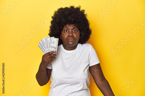African-American woman holding cash, displaying wealth shrugs shoulders and open eyes confused.