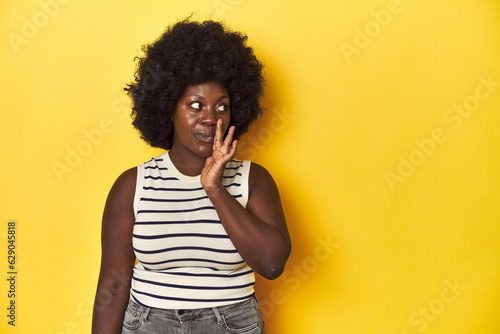 African-American woman with afro, studio yellow background is saying a secret hot braking news and looking aside © Asier