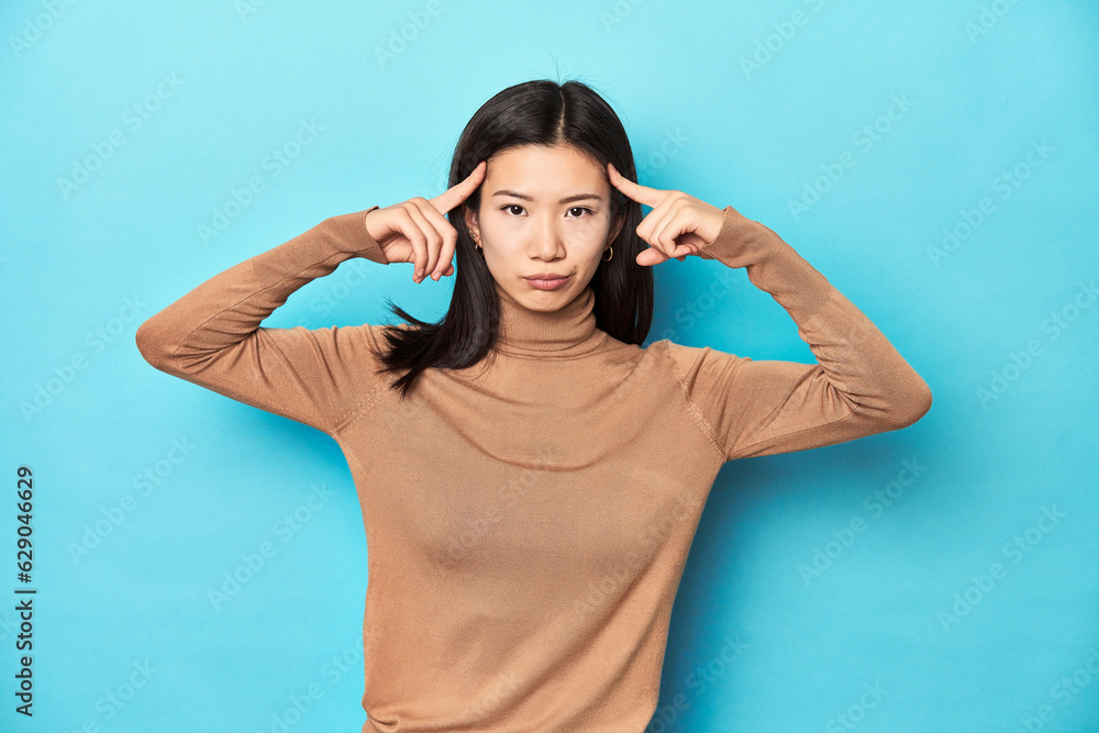 Young Asian woman in brown turtleneck, focused on a task, keeping forefingers pointing head. Young Asian woman in brown turtleneck, focused on a task, keeping forefingers pointing head.