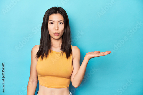 Asian woman in summer yellow top, studio setup, impressed holding copy space on palm.