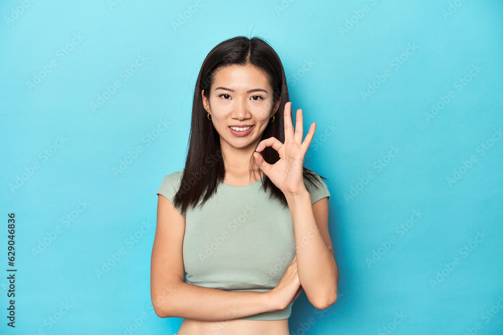 Asian woman in summer green top, studio backdrop, winks an eye and holds an okay gesture with hand. Asian woman in summer green top, studio backdrop, winks an eye and holds an okay gesture with hand.