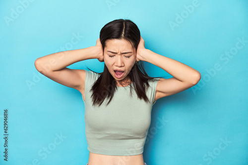 Asian woman in summer green top, studio backdrop, covering ears with hands trying not to hear too loud sound.