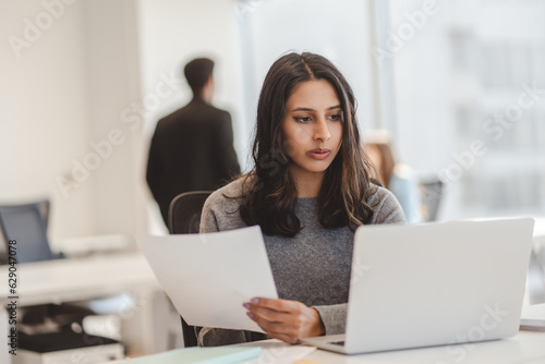 Serious young indian woman, entrepreneur holding documents while working on laptop