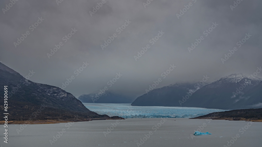 Obraz premium The blue glacier of Perito Moreno stretches between the mountain slopes. Breakaway icebergs float in the lake. Cloudy and foggy. Argentina. El Calafate. Lago Argentino.