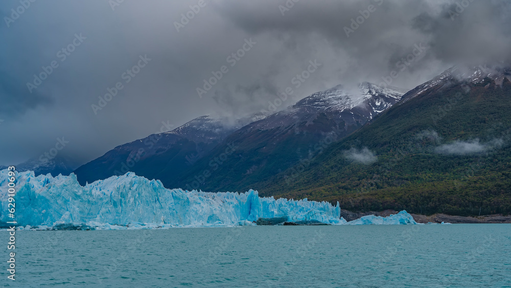 The amazing Perito Moreno glacier. A wall of cracked blue ice rises above a turquoise lake. The peaks of the snow-capped mountains hide in the clouds. Argentina. El Calafate