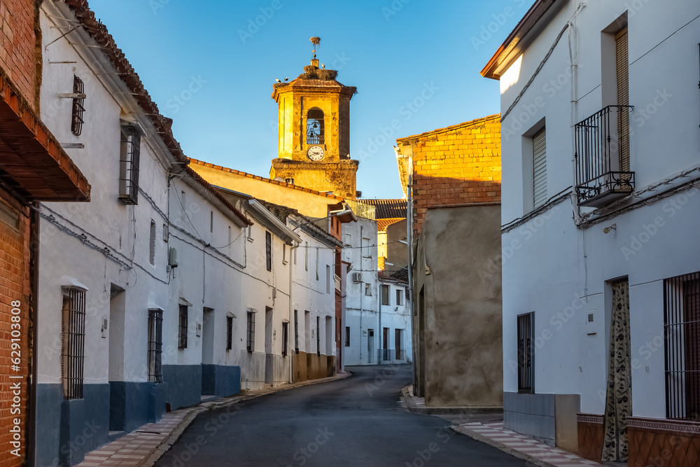 Alley with old houses painted white and tower of the Catholic church illuminated by the light of dawn, Agudo, Ciudad Real.