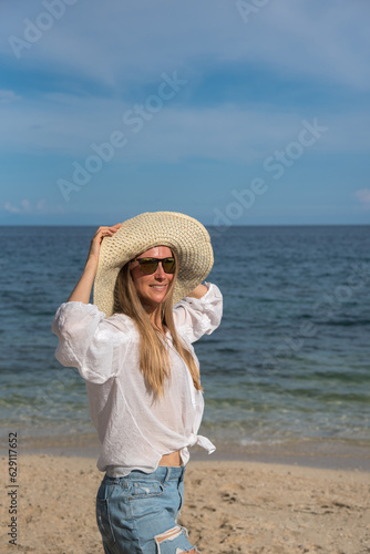 A blond woman with sunglasses is standing by the sea. She is touching her straw hat. A girl with long hair and a white shirt spends her vacation on the beach. Holidays and travel to other countries