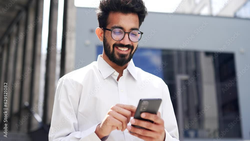 Close up. A young smiling businessman is using a smartphone while standing on the street near an office building. Happy handsome bearded male checking email, chatting online, texting with a client Close up. A young smiling businessman is using a smartphone while standing on the street near an office building. Happy handsome bearded male checking email, chatting online, texting with a client