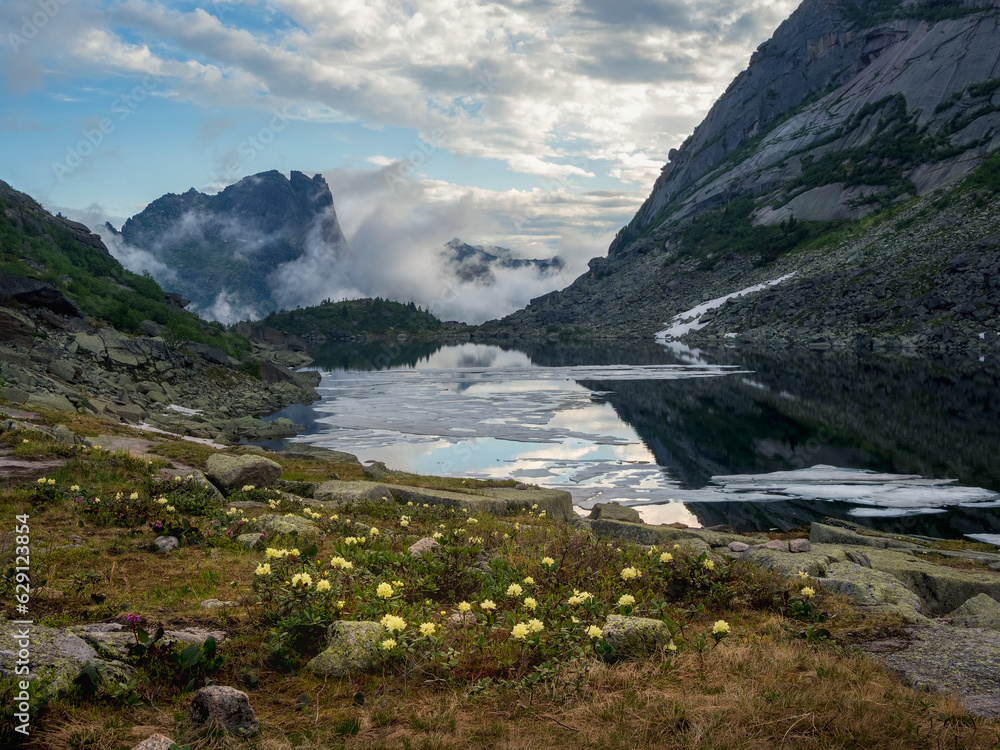 Amazing morning view of the summer mountain, fog over the mountain slopes in the distance, white clouds filling the mountain gorge. A clear ice blue mountain lake and a bright flower meadow.
