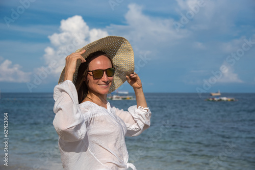 A happy blond woman with sunglasses is standing by the sea. She is touching her straw hat. A girl with long hair and a white shirt spends her vacation on the beach. Holidays to other countries