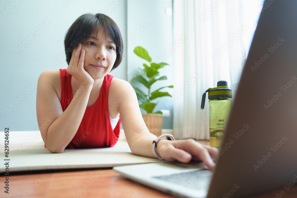 Obraz premium Asian chinese woman in sportwear lying on the floor using Laptop while doing yoga exercise at home. Typing on computer.