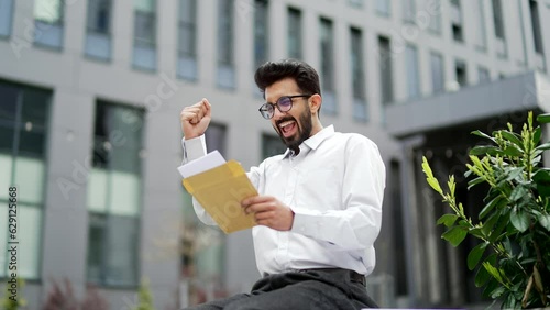 Happy excited young businessman reading letter with great news while standing on the street near an office building. A man is celebrating success, happy to receive a pleasant notification. Wow effect