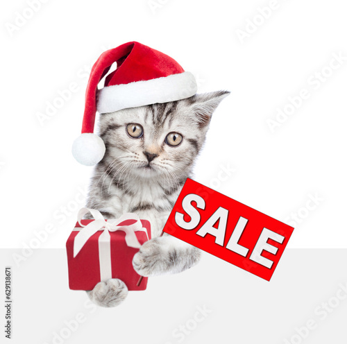 Tabby kitten wearing red christmas hat holds gift box and shows signboard with labeled "sale"above empty white banner. isolated on white background