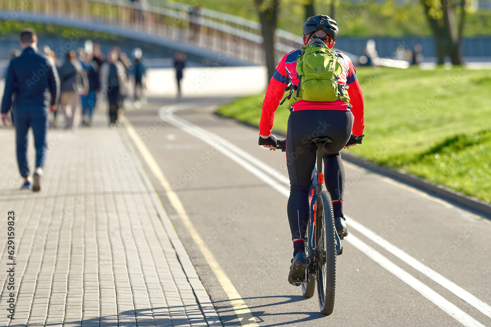 Fototapeta premium Man in sport outfits cycling. Cyclist in sport protective gear riding on bike lane in the city. Cyclist commutes on bike. Man riding bike in city park.