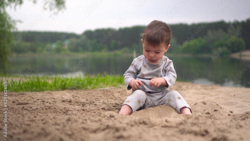 A little boy sits on the beach by the river and plays in the sand. A two-year-old child plays in the sand against the background of the river. A little boy sits on the beach by the river and plays in the sand. A two-year-old child plays in the sand against the background of the river.