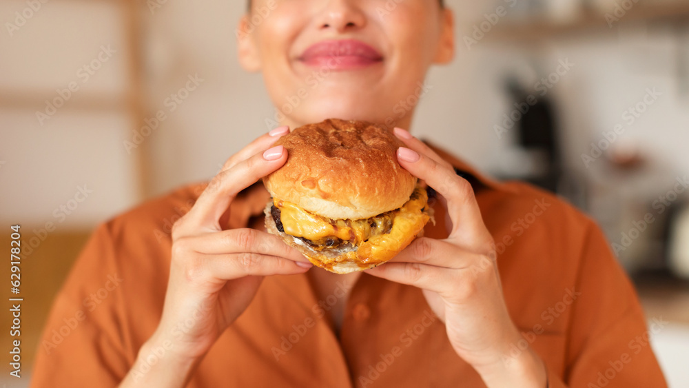 Closeup shot of young woman in casual clothes holding american classic fast food burger, sitting in kitchen, panorama Closeup shot of young woman in casual clothes holding american classic fast food burger, sitting in kitchen, panorama