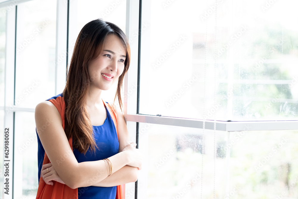 Beautiful Asian woman standing and smilling against the glass window. Young lady looking outside the window. Beautiful Asian woman standing and smilling against the glass window. Young lady looking outside the window.