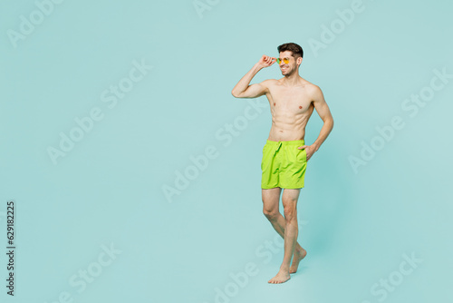Full body side view young sexy man wear green shorts swimsuit sunglasses relax near hotel pool look aside on area mock up isolated on plain blue background. Summer vacation sea rest sun tan concept.