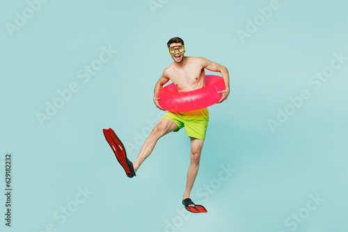 Full body satisfied young man wear green shorts swimsuit relax near hotel pool hold in hand inflatable rubber ring flippers isolated on plain blue background. Summer vacation sea rest sun tan concept. photo