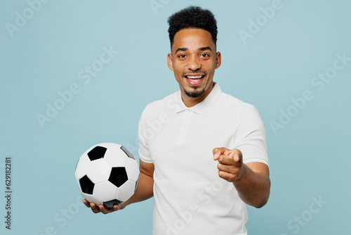 Young man fan wears t-shirt point index finger camera on you cheer up support football sport team hold in hand soccer ball watch tv live stream isolated on plain pastel blue color background studio.