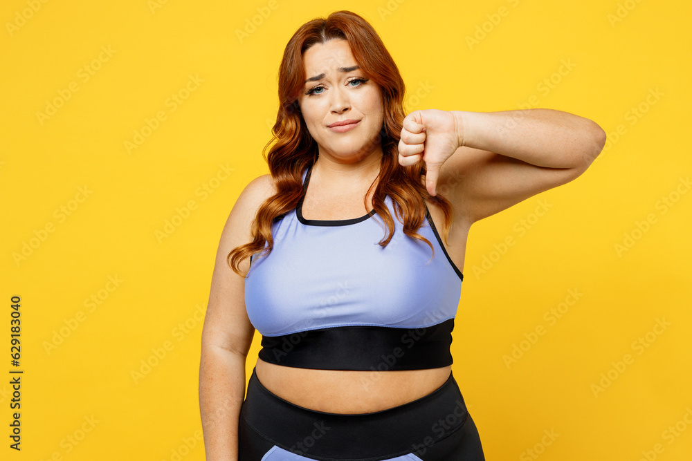 Young sad chubby overweight plus size big fat fit woman wears blue top warm up training showing thumb down dislike gesture isolated on plain yellow background studio home gym. Workout sport concept. Young sad chubby overweight plus size big fat fit woman wears blue top warm up training showing thumb down dislike gesture isolated on plain yellow background studio home gym. Workout sport concept.