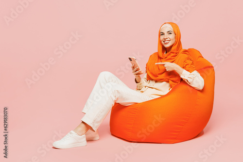 Full body young arabian asian muslim woman wearing orange abaya hijab sit in bag chair hold use point finger on mobile cell phone isolated on plain pink background. Uae middle eastern islam concept.
