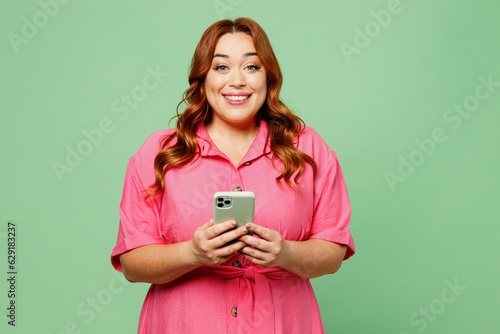 Young happy chubby overweight redhead woman wear casual clothes pink dress hold in hand use mobile cell phone chatting online isolated on plain pastel light green color background. Lifesyle concept.
