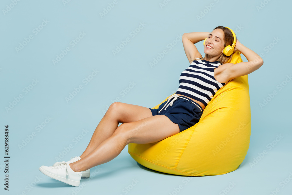 Obraz premium Full body young woman she wear striped tank shirt casual clothes sit in bag chair listen to muisc in headphones close eyes isolated on plain pastel light blue cyan background studio Lifestyle concept