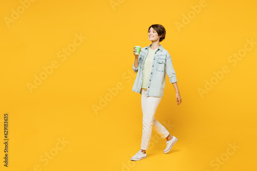 Full body young happy woman she wear green t-shirt denim shirt casual clothes hold takeaway delivery craft paper brown cup coffee to go isolated on plain yellow background studio. Lifestyle concept.