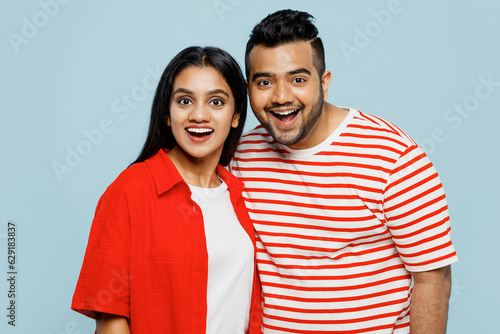 Young surprised shocked cheerful couple two friends family Indian man woman wear red casual clothes t-shirts together loking camera, joiking isolated on pastel plain light blue cyan color background.