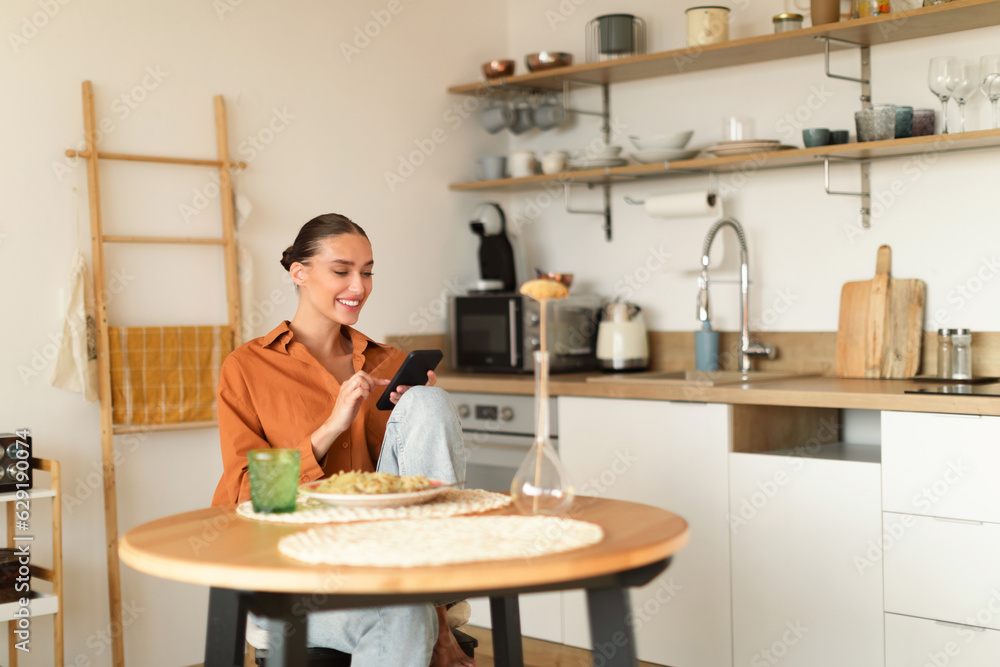 Cheerful young woman sitting in kitchen, using smartphone while having dinner, copy space Cheerful young woman sitting in kitchen, using smartphone while having dinner, copy space