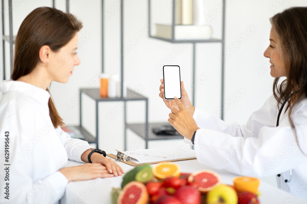 Happy european middle aged lady doctor nutritionist in white coat advises woman patient, recommends app on phone