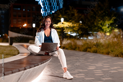 Female freelancer working on laptop  sitting on bench at night in city park on modern urban background  free space