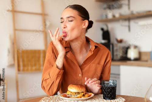Contented caucasian lady licking her fingers while eating delicious french fries and hamburger, kitchen interior
