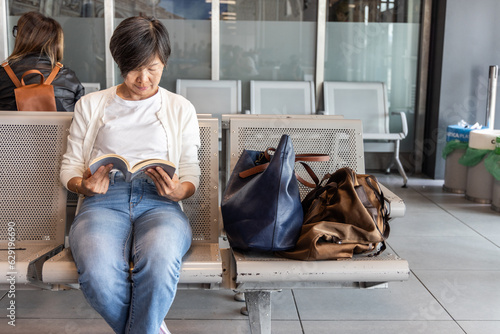 Middle age Asian woman with luggage reading book while waiting for departure in airport terminal