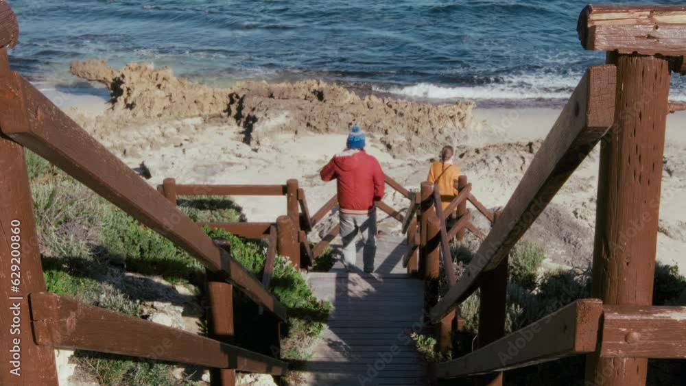 Rear view of couple of hikers coming down to ocean coast through a picturesque bushy landscape and wooden stairs. Male and female travelers navigate their way down a scenic trail, surrounded by lush Rear view of couple of hikers coming down to ocean coast through a picturesque bushy landscape and wooden stairs. Male and female travelers navigate their way down a scenic trail, surrounded by lush
