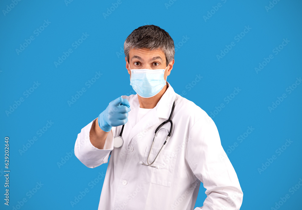 Doctor in facial mask pointing at camera over blue background Doctor in facial mask pointing at camera over blue background