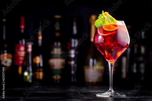 Red sangria summer alcoholic drink with red spanish wine, peach, plum, orange, lime and ice. Black bar counter background, steel bar tools and bottles