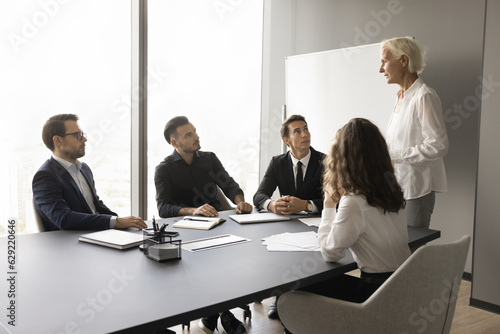 Mature company mentor, coach training employees, giving consultation, advice. Business team leader standing at meeting table, speaking to sitting, listening younger colleagues